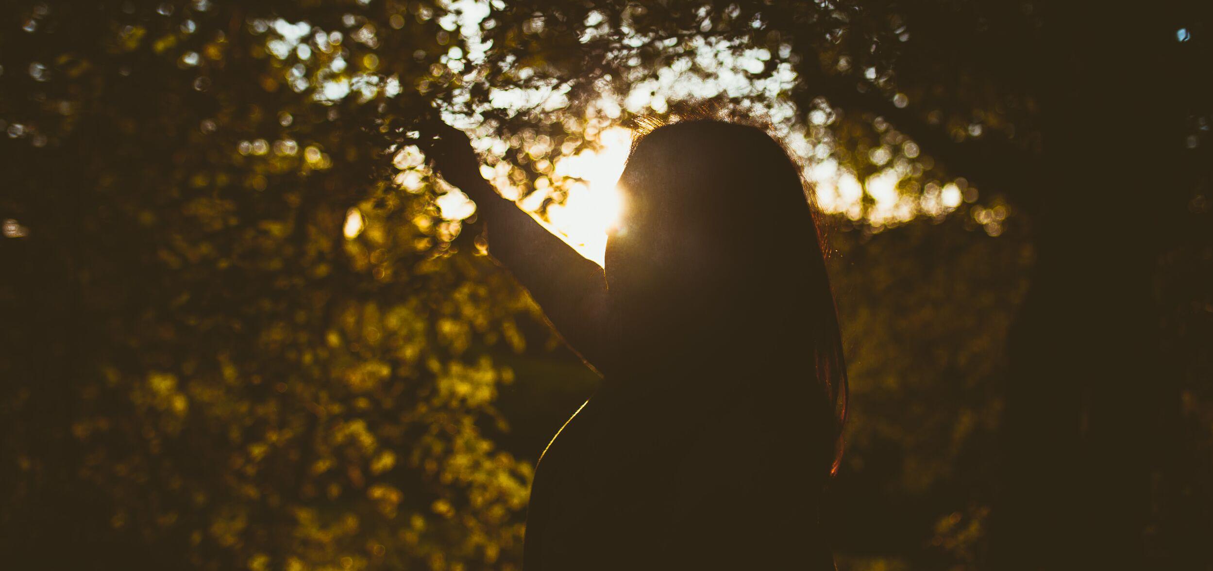 A woman's silhouette standing behind trees with her arm outstretched and sunlight seeping through. 