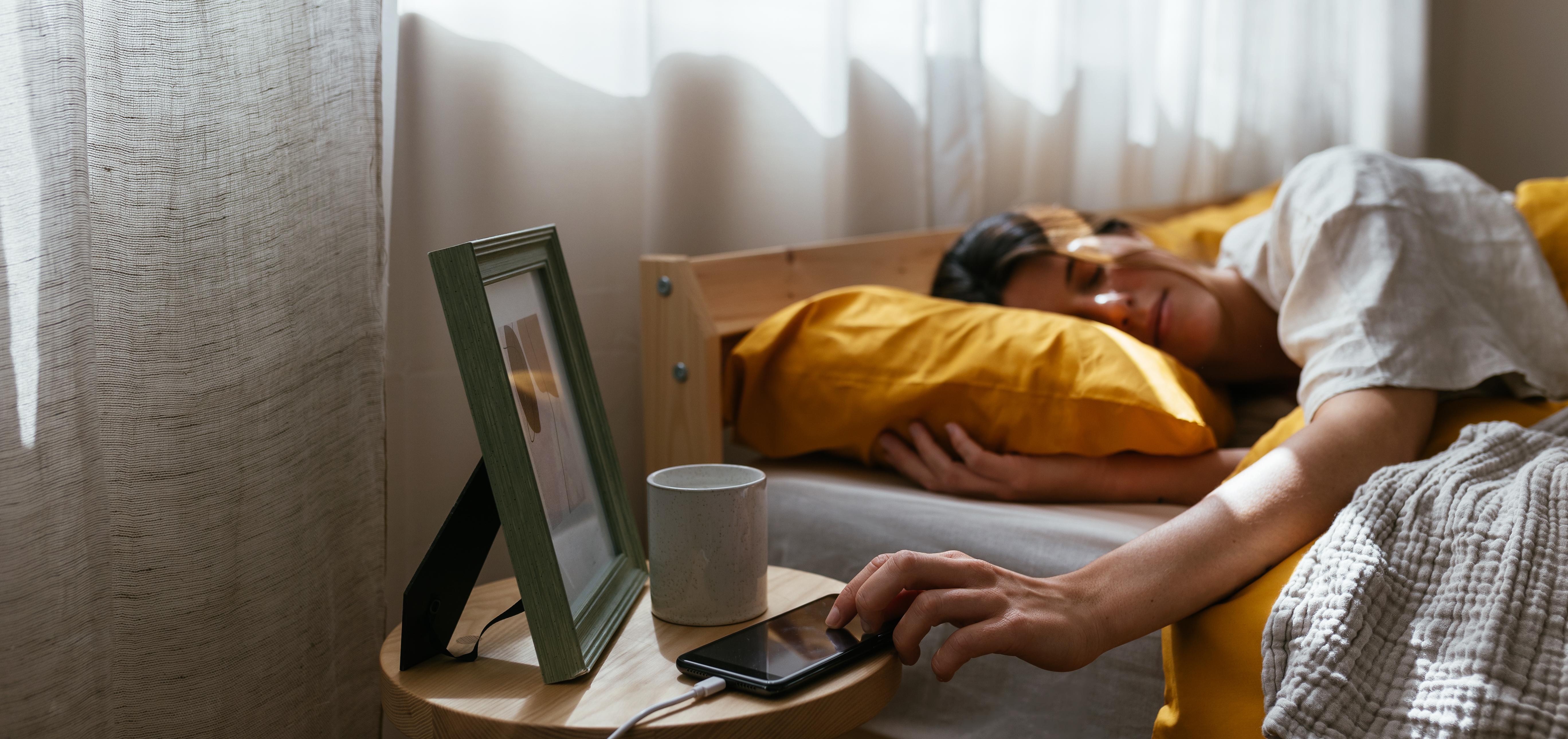 A woman in bed reaching for her phone as sun peeps through her window