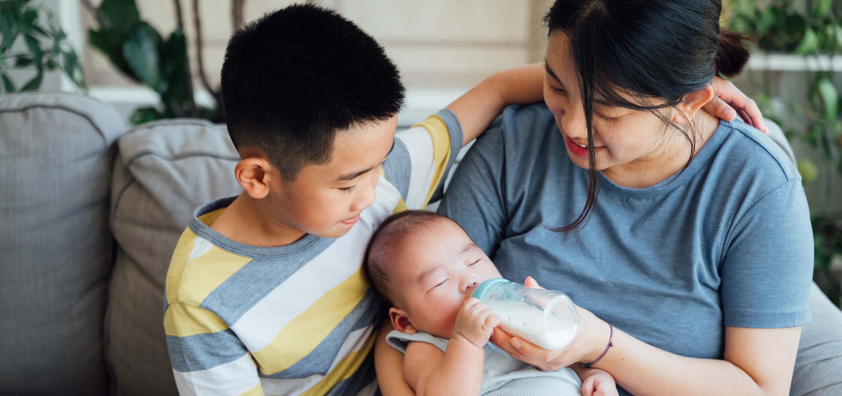 mother feeding her baby with a bottle of milk while young son looks on