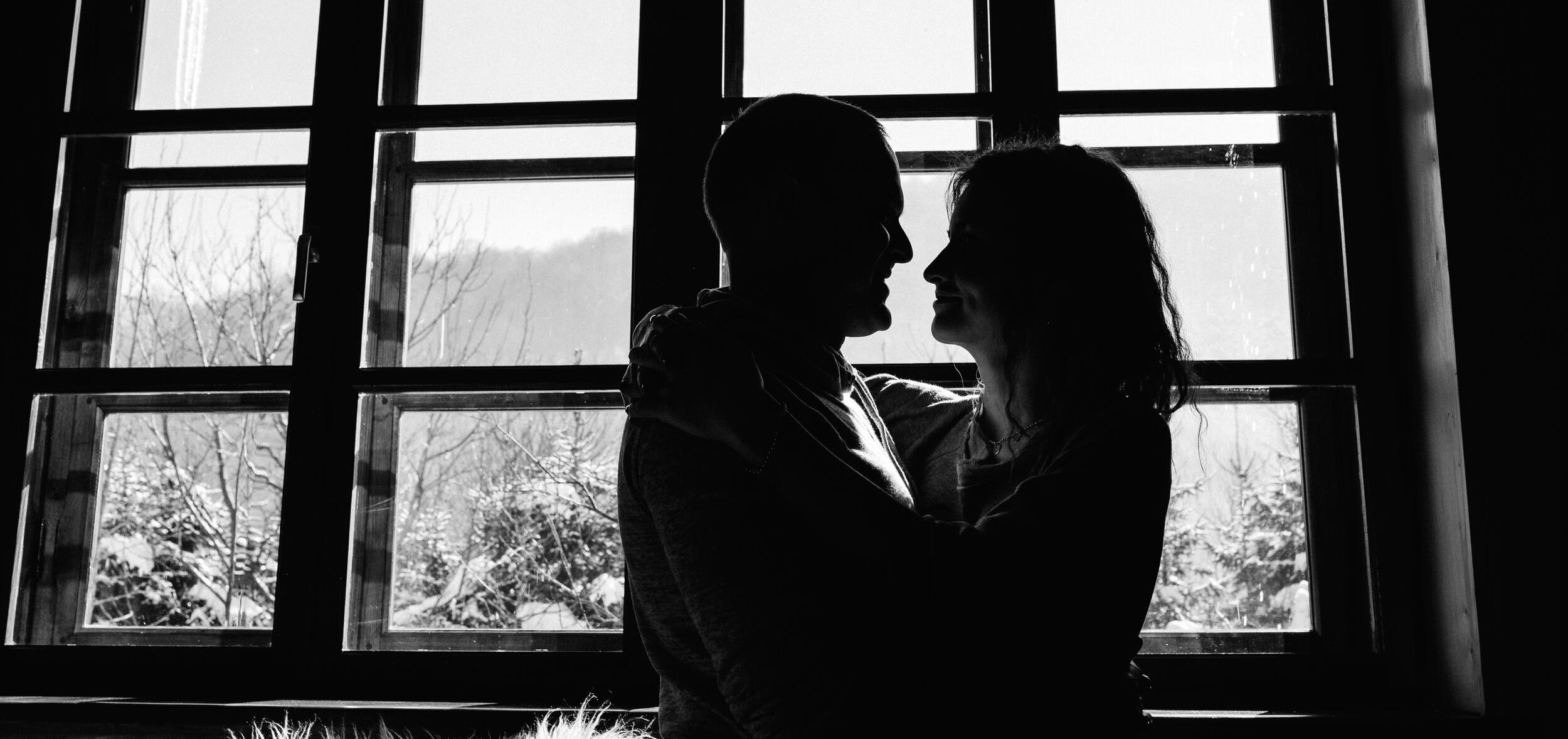 In black and white, a couple embraces in front of a large window