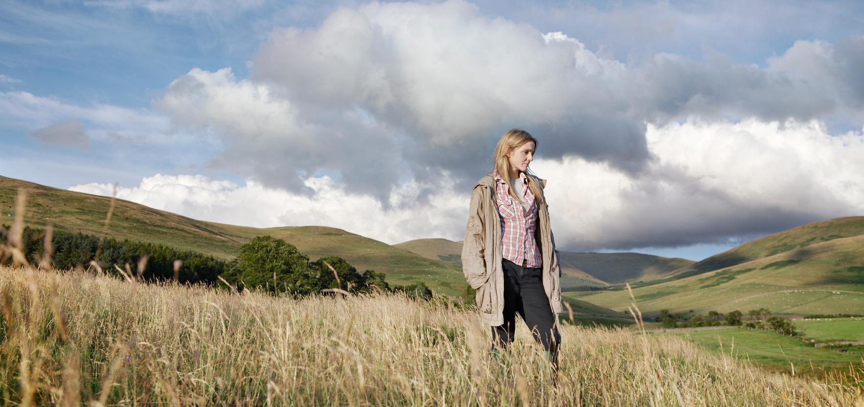 Woman with rolling hills behind her, looking pensive. 