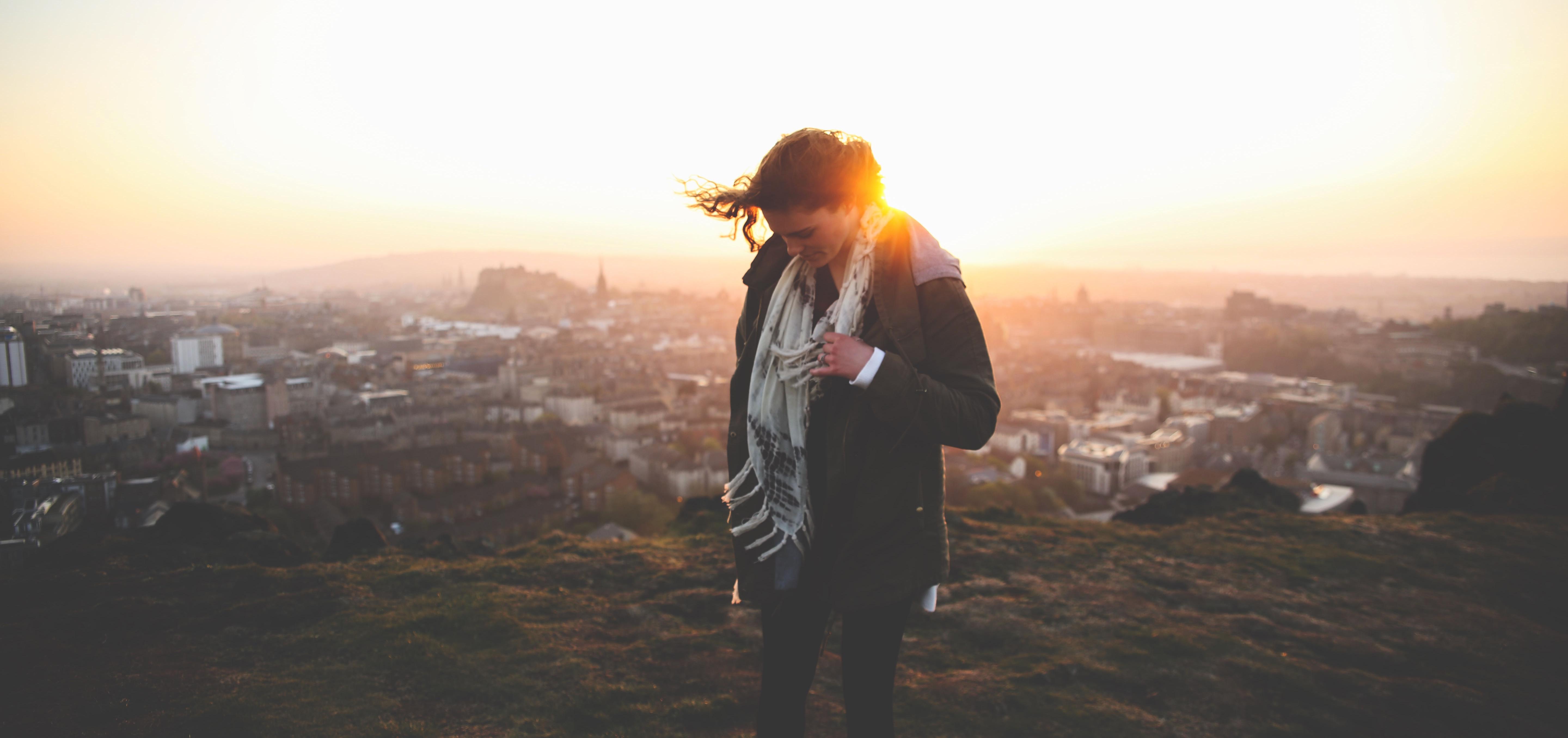 Woman with jacket and scarf stands on a breezy hilltop overlooking a city at sunset