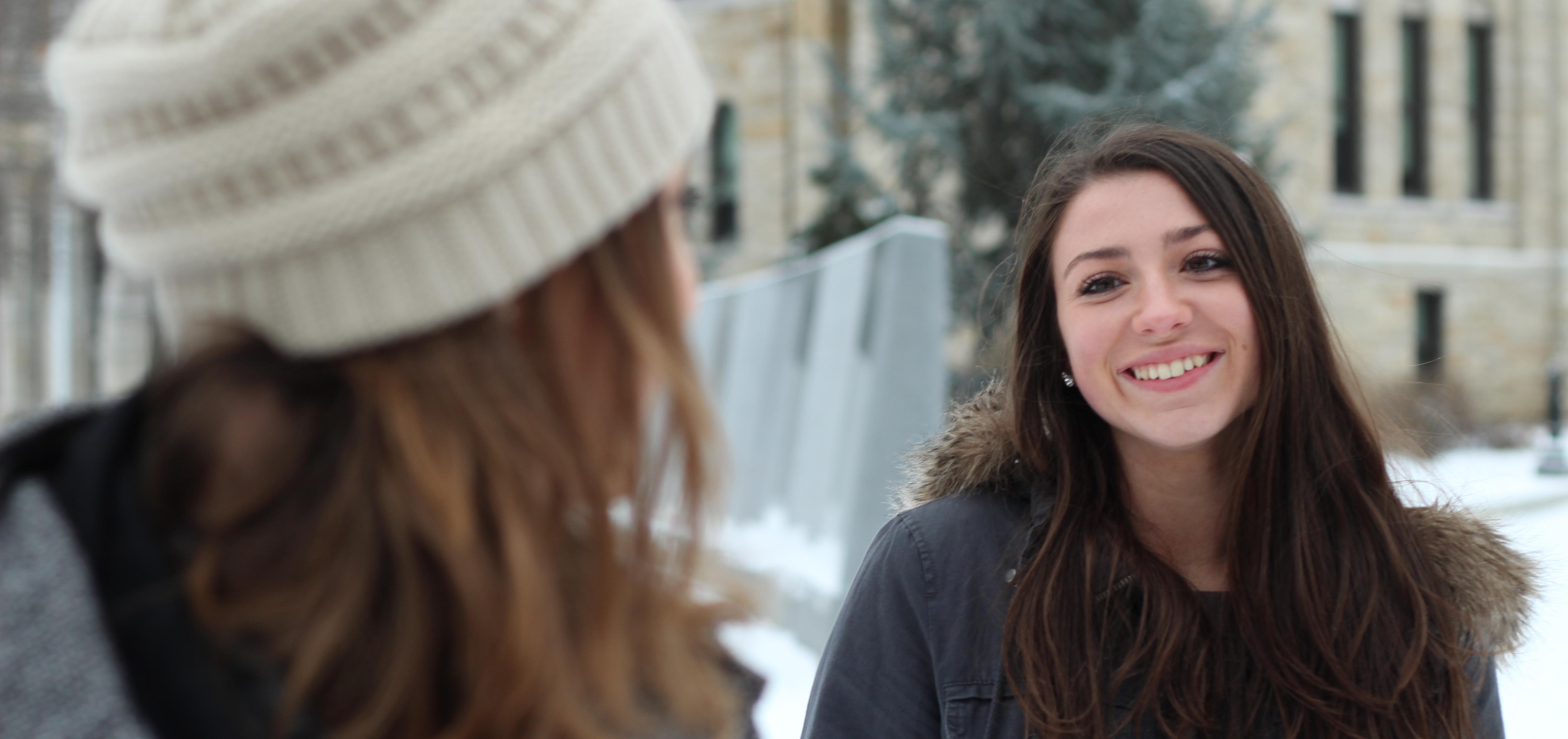 A young woman in a winter jacket smiles at another woman in a white hat.