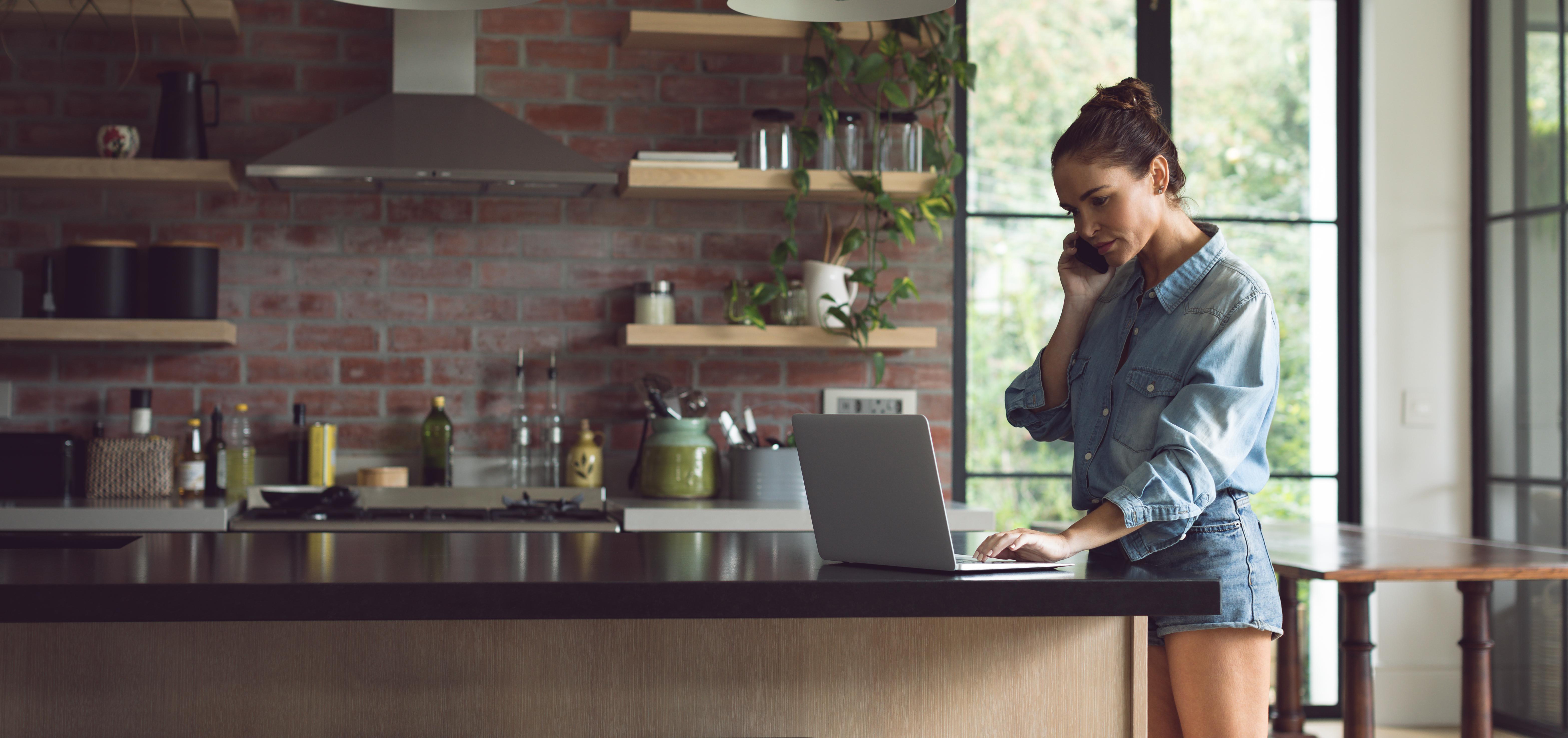 A woman standing at her kitchen counter using her laptop while talking on the phone