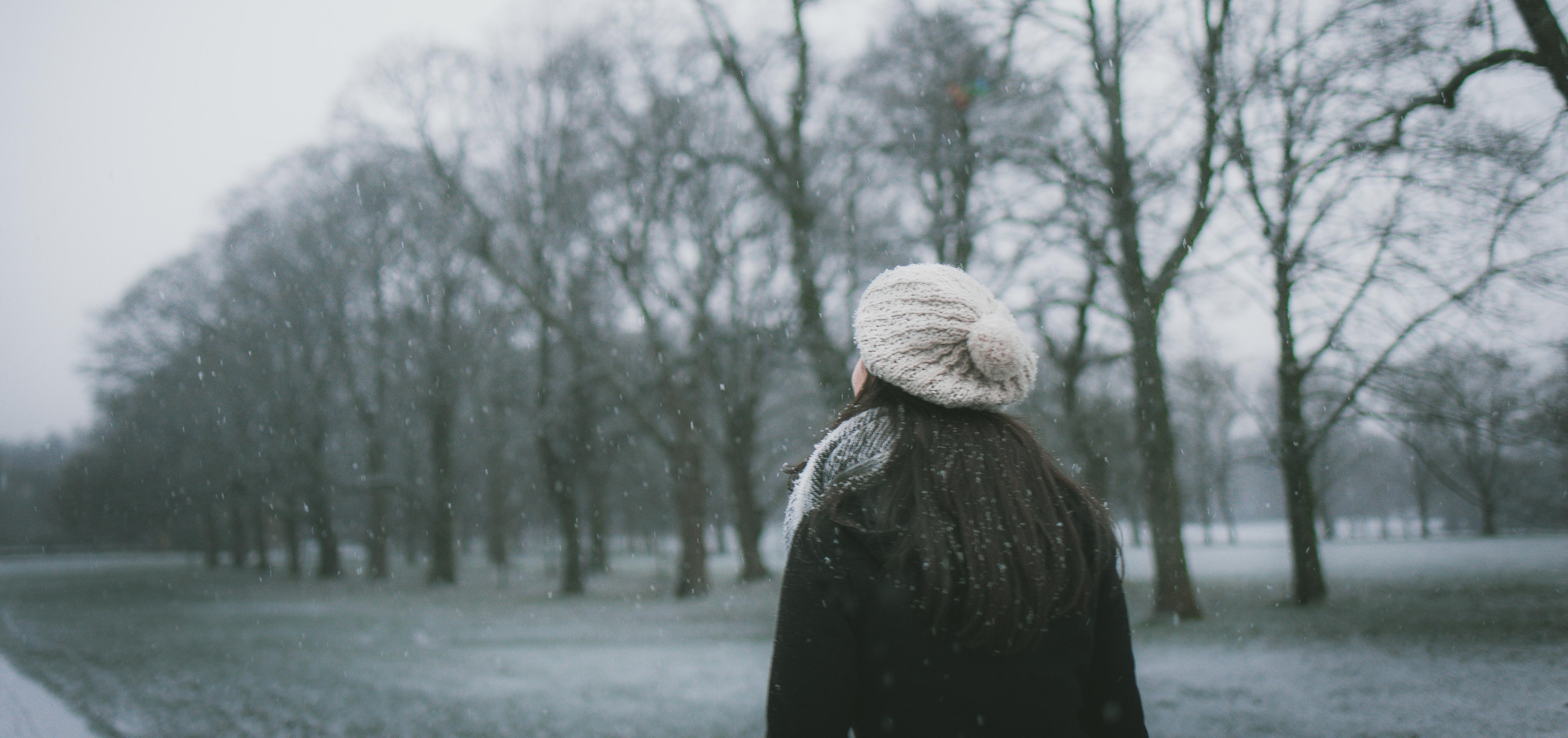 A woman with long hair wearing a white beanie walking through a snowy park with trees