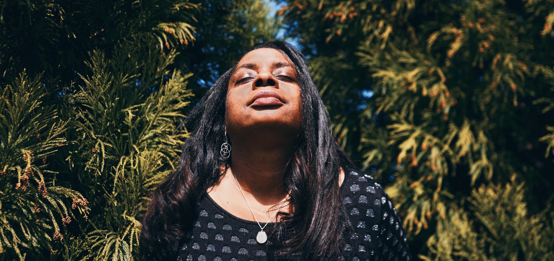 woman standing among trees, her face turned up toward the sunlight with eyes closed gently